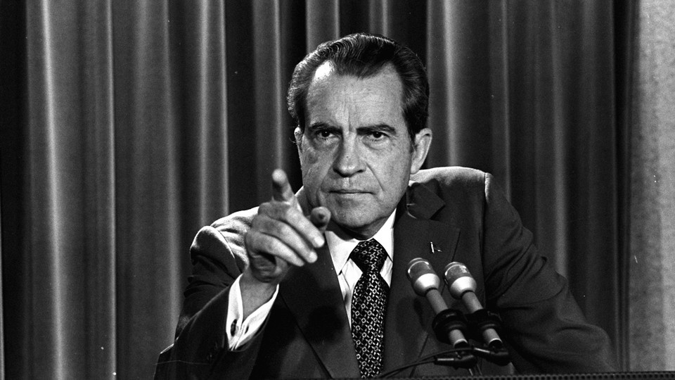 Black-and-white photo of Richard Nixon standing at a lectern with two microphones, facing the camera and pointing his finger at something or someone in the near distance