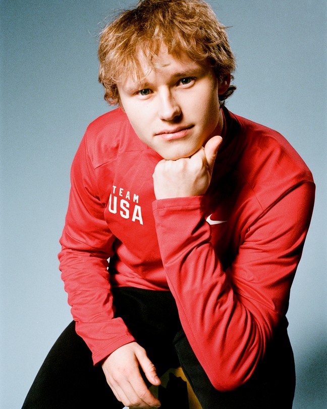 photo portrait of young man with red hair in red long-sleeve TEAM USA shirt and black pants, sitting on stool with one hand under chin