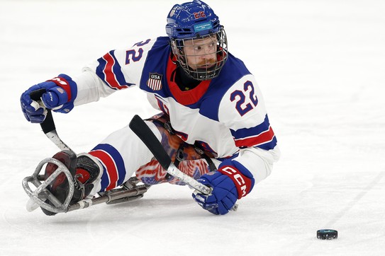 A hockey player, seated in an ice-hockey sled, maneuvers with a puck during a game.