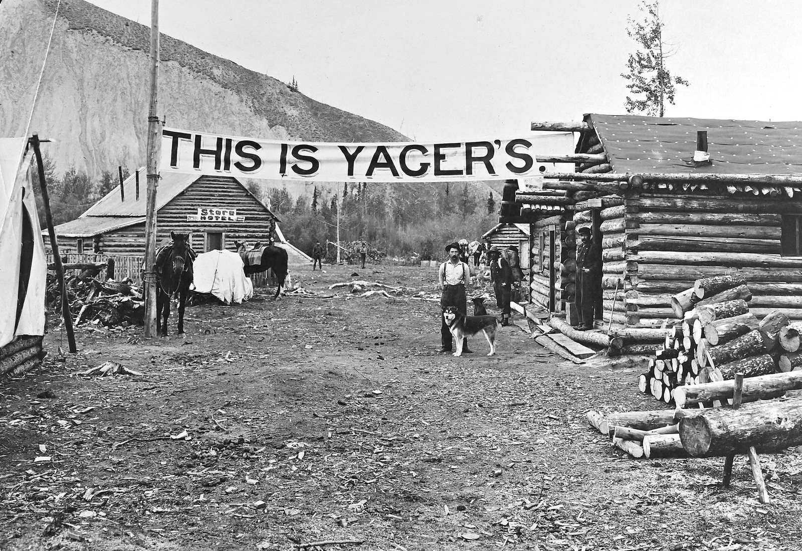 People gather outside a combination roadhouse, store, and hotel, consisting of several log buildings, under a banner reading
