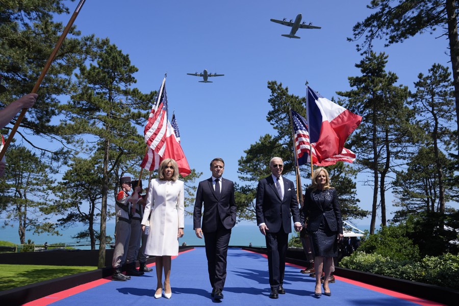The presidents of France and the United States, and their spouses, walk past soldiers holding American and French flags, as several large military aircraft fly overhead.
