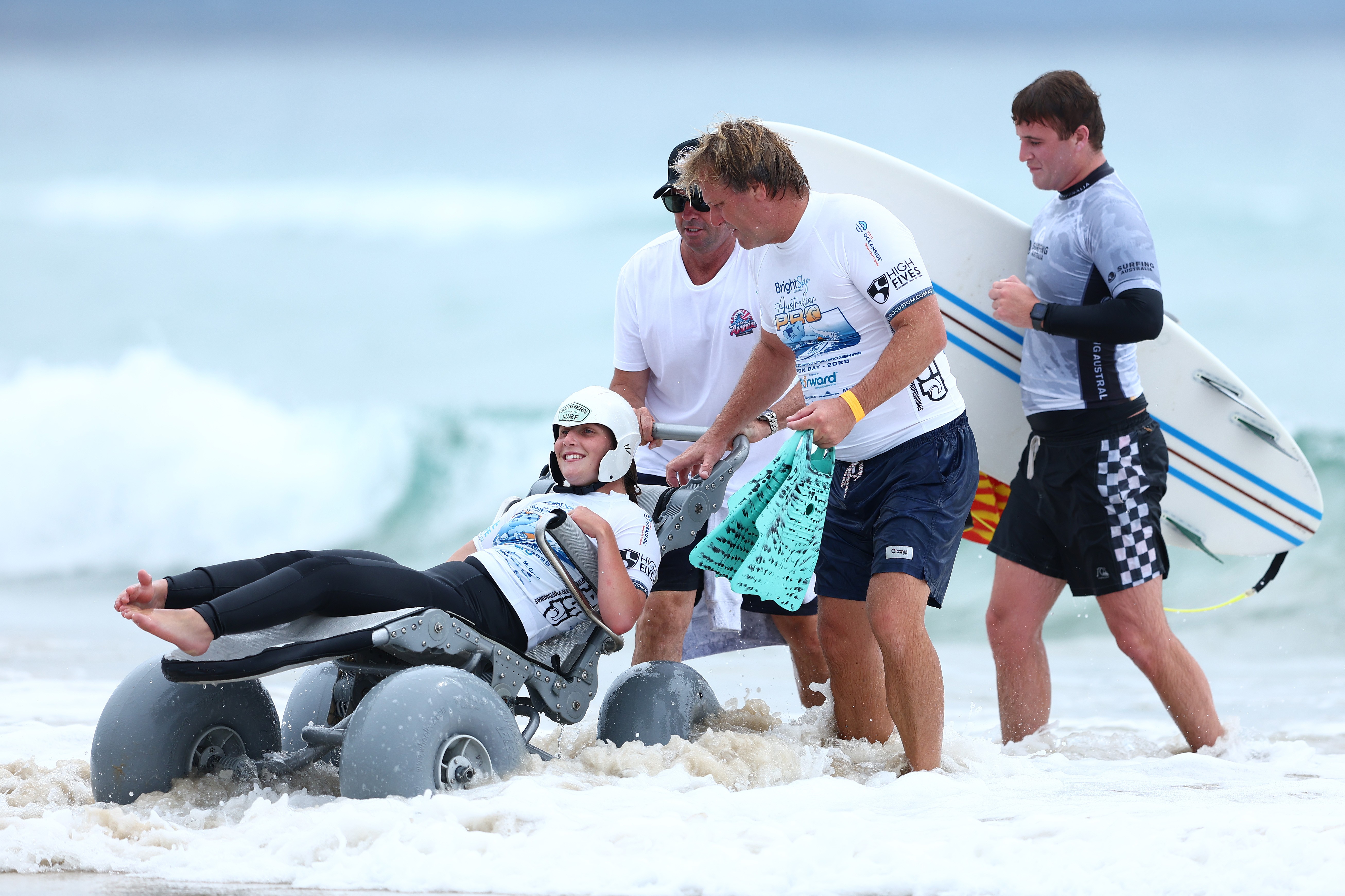 A para surfer is assisted by others as they push her out of the surf on a wheelchair with large rounded wheels.