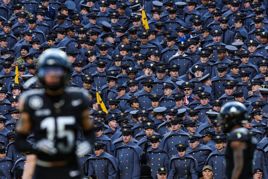 Dozens of cadets in dress uniforms stand in stadium seating, watching football players in the foreground.