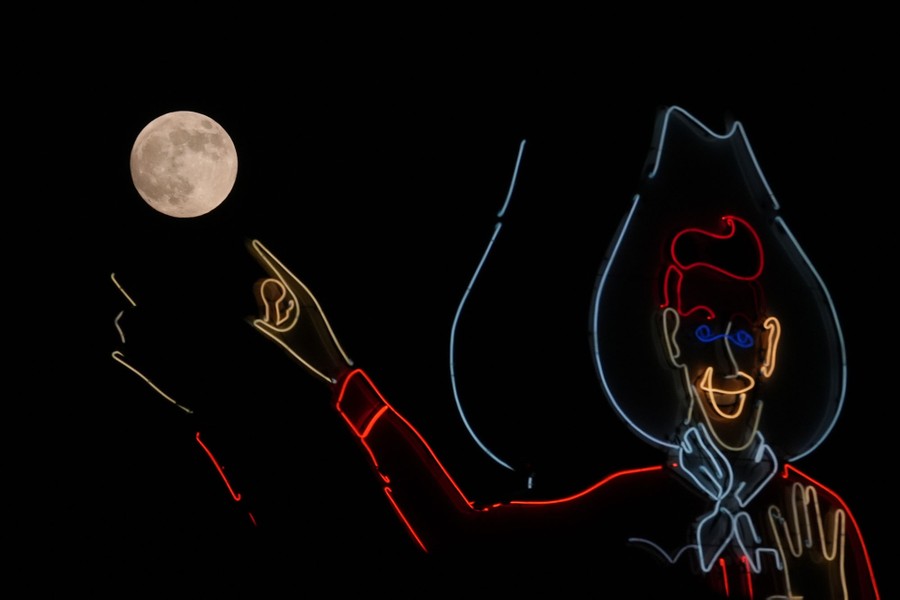 The full moon rises beyond a large neon cowboy sign, which appears to point at the moon.