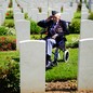 An elderly veteran wearing many medals sits in a wheelchair, saluting, among many rows of headstones in a cemetery.