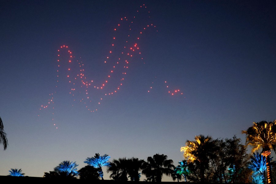 Groups of flying drones form swooping dotted lines in the sky above palm trees.