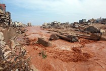 Muddy water rushes in a river channel past heavily damaged buildings after a flood.