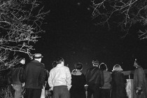 A black-and-white photograph of a group of people looking up at the night sky, which is framed by some trees