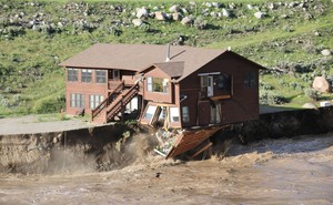 A fast-flowing river undercuts its banks, causing a two-story wooden cabin to start collapsing.