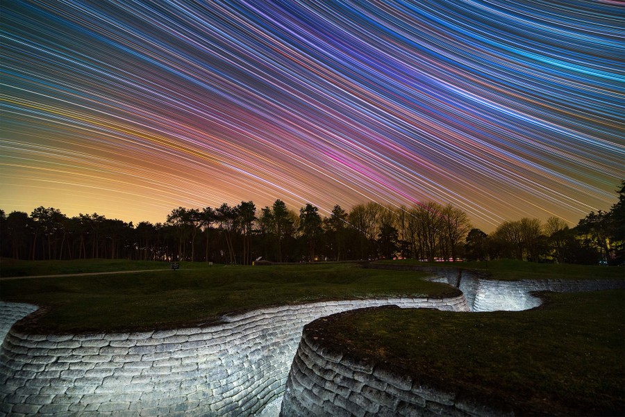 A long-exposure photo shows the stars as many bright streaks across the night sky, above preserved trenches in a memorial park.