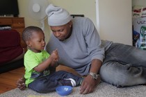 A father sits with his son on the carpet and looks at him lovingly while the child stares at a snack.