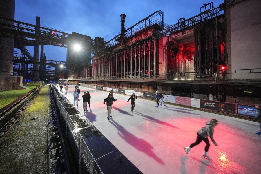 People ice-skate on a long rink situated among old industrial buildings.