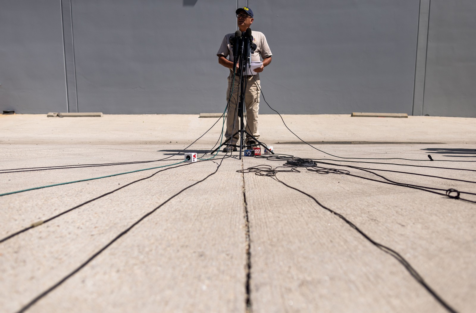 A person answers questions during a press event, standing in front of a microphone stand in a parking lot, seen from a low angle.