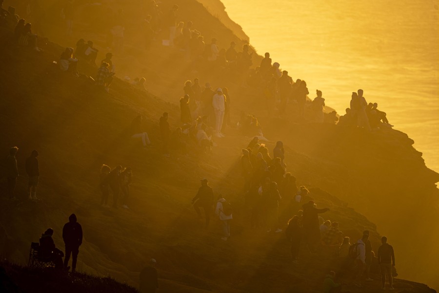 A crowd of spectators, bathed in golden light, watches surfers from a steep coastal hillside.