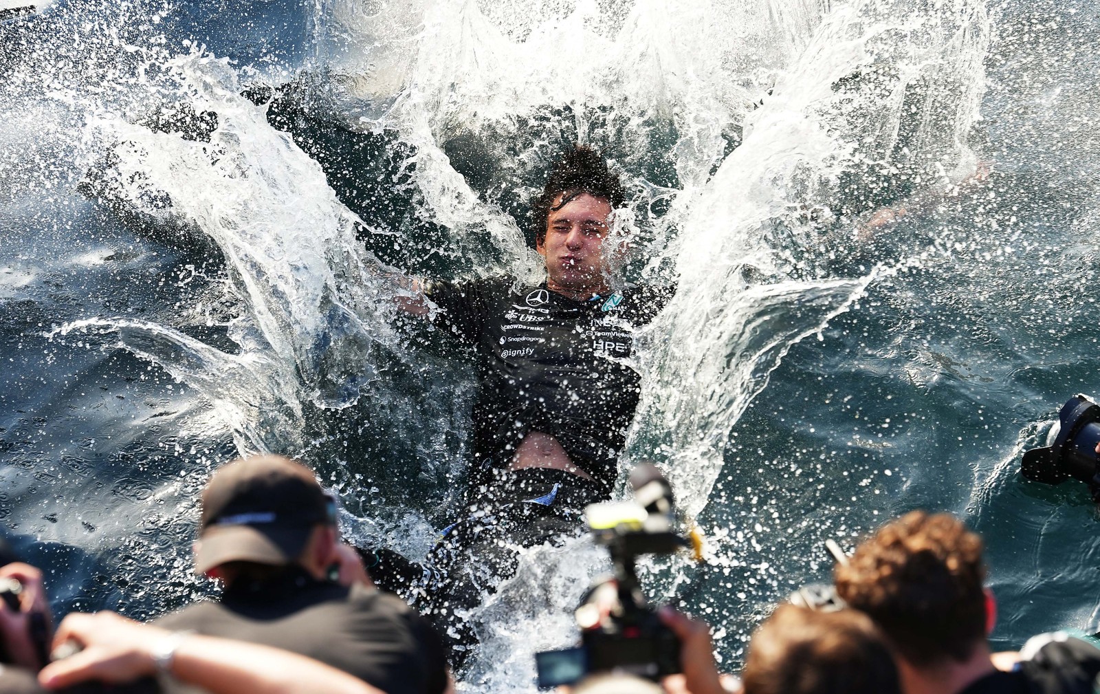 A race car driver makes a splash after jumping into a harbor in celebration, as others take photographs.
