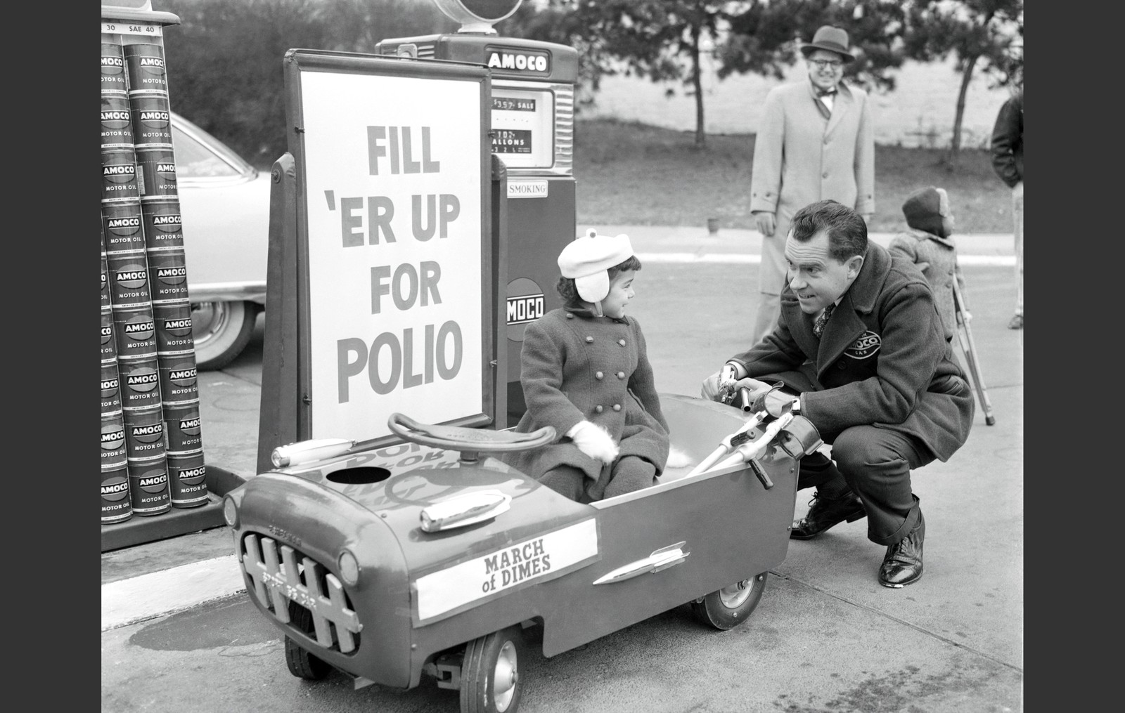 Vice President Richard Nixon, wearing a jacket belonging to an Amoco gas station, kneels down behind a child's small pedal car, pretending to fill it with gasoline, as the child driver looks back at him. They are in front of a sign that reads 'Fill 'er up for polio.'