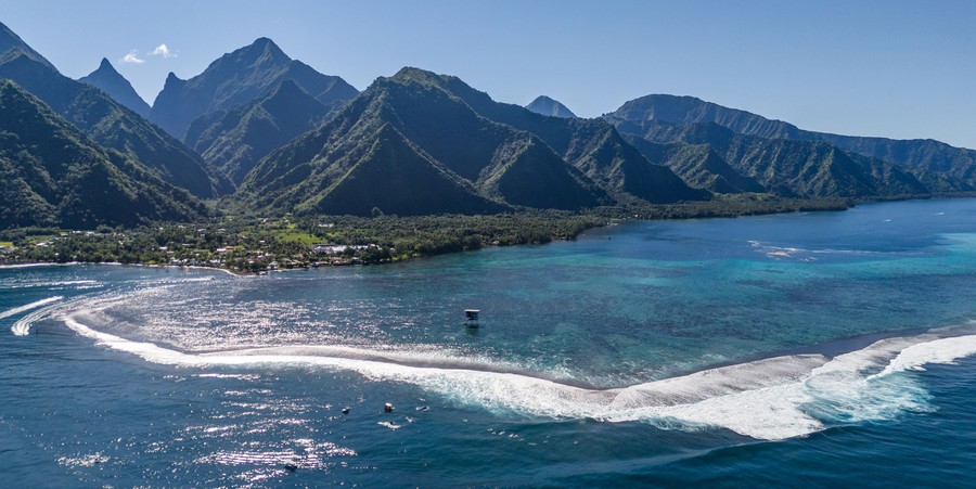 An aerial view of a tropical shoreline featuring steep mountains and crashing waves