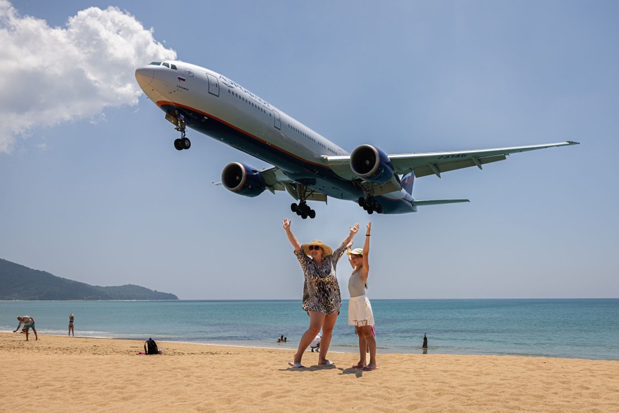 Two people pose on a beach as a large passenger aircraft flies closely above, on its way to a landing.