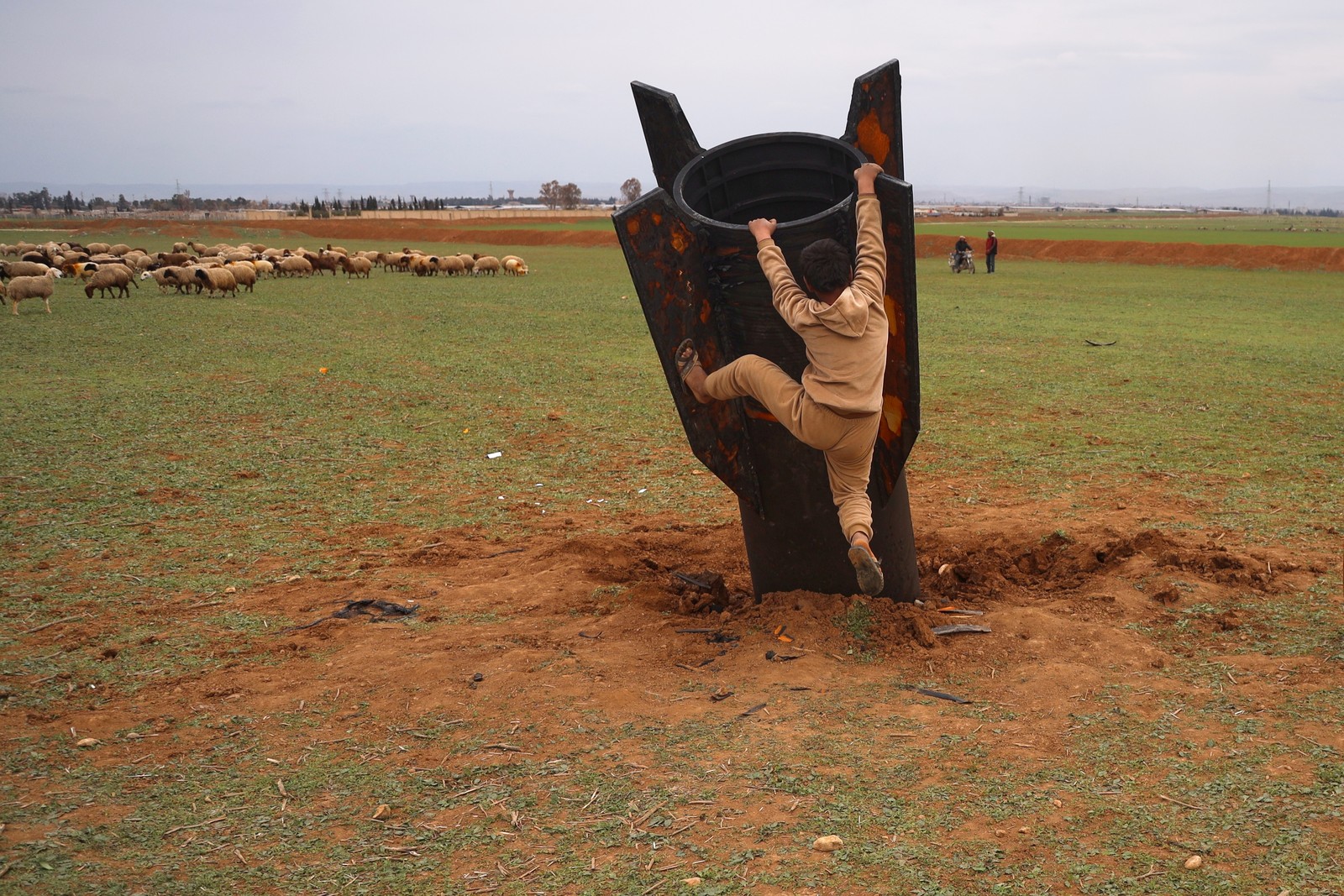 A boy tries to climb on an unexploded missile that landed in an open field, its nose embedded in the dirt.