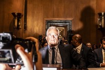 A color photo of RFK Jr. in a suit, in a wood-paneled room, surrounded by other people in suits and lit dramatically by a camera flash