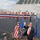 Trump waves in front of flags and a large military ship filled with men taking photos of him