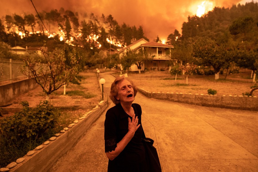 A woman reacts, hand to her chest, standing in a driveway, as a forest fire rages behind her house.