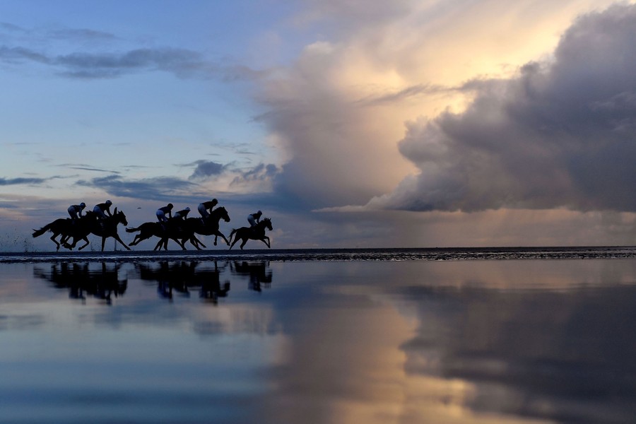 Several racers ride horses along a beach, with large clouds in the background.