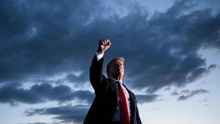 President Trump, standing, raises his fist, with a dark sky behind him