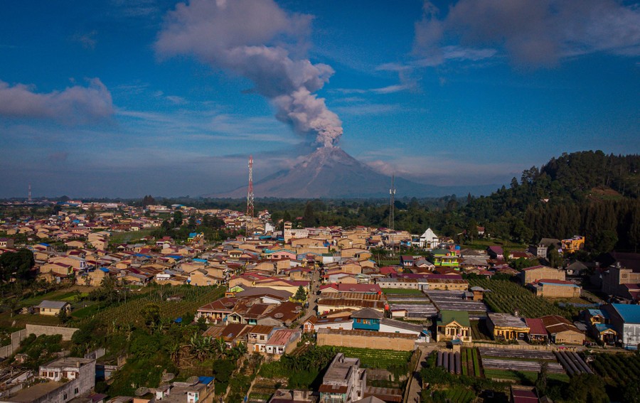 A volcano erupts, seen beyond a town in the foreground.