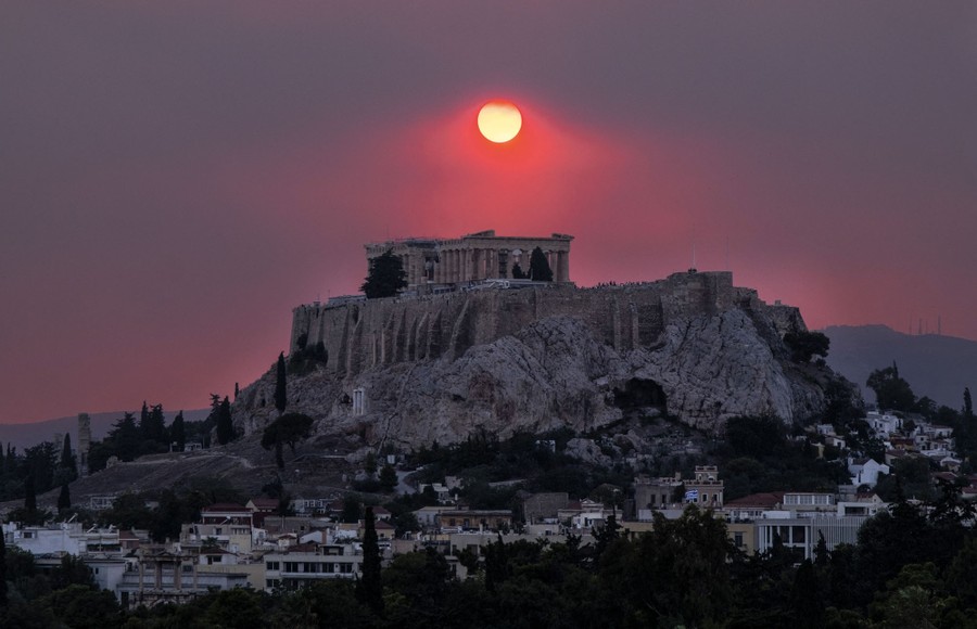 The ancient ruin of the Parthenon sits atop a steep hill, seen beneath a smoke-filled sky at sunset