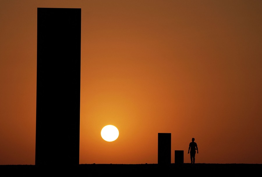 The silhouette of a sculpture, consisting of several tall obelisks, with the setting sun behind it against a rust-colored sky