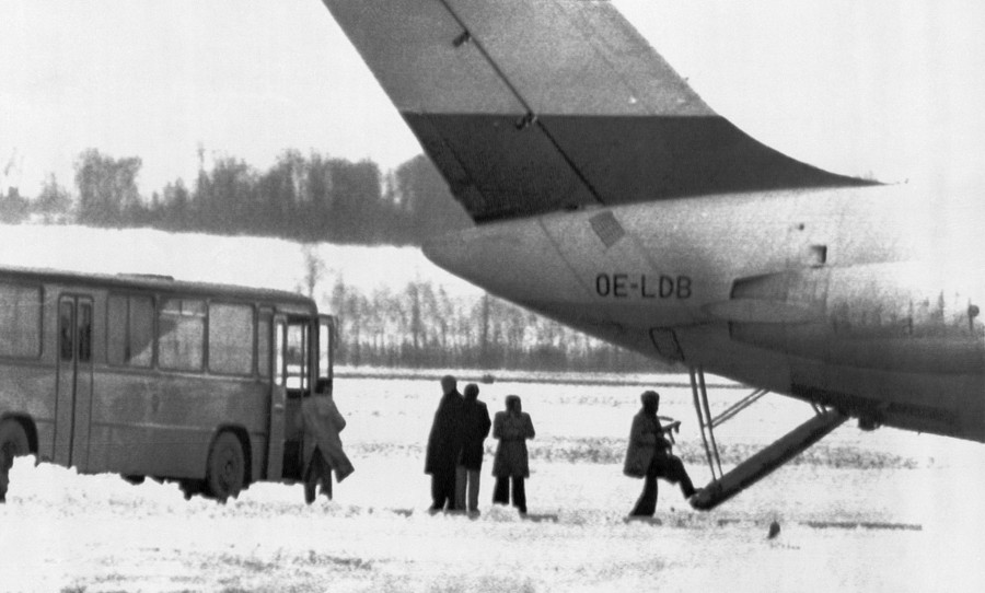 Several people stand outside a large aircraft, as one (carrying a weapon) boards through a rear staircase.