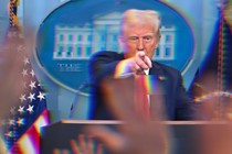 A blurred color photograph of Donald Trump in a White House press conference, pointing forward over a podium.