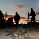 A photograph with camo gear and a rifle illuminated in the foreground and a half-dozen soldiers silhouetted in the background.