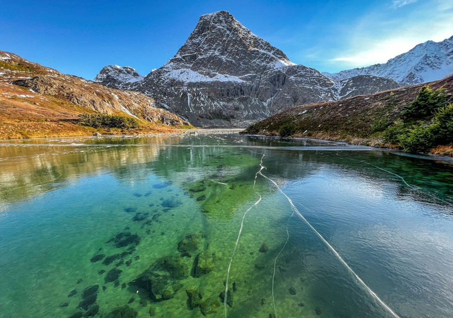 Green water can be seen in an alpine like, through its frozen surface, with a tall mountain nearby.