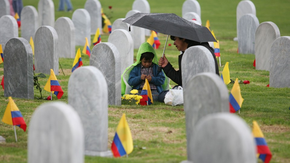 A child sits under an umbrella with an adult in a cemetery. Colombian flags are in the ground next to each headstone.