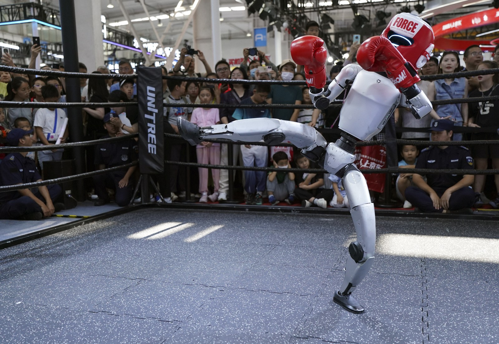 A small humanoid robot wearing boxing gloves performs a kick in front of an audience.