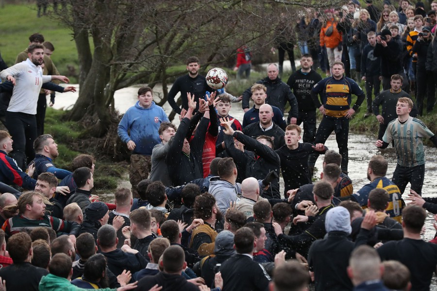 Dozens of men compete to get their hands on a ball, near a river.