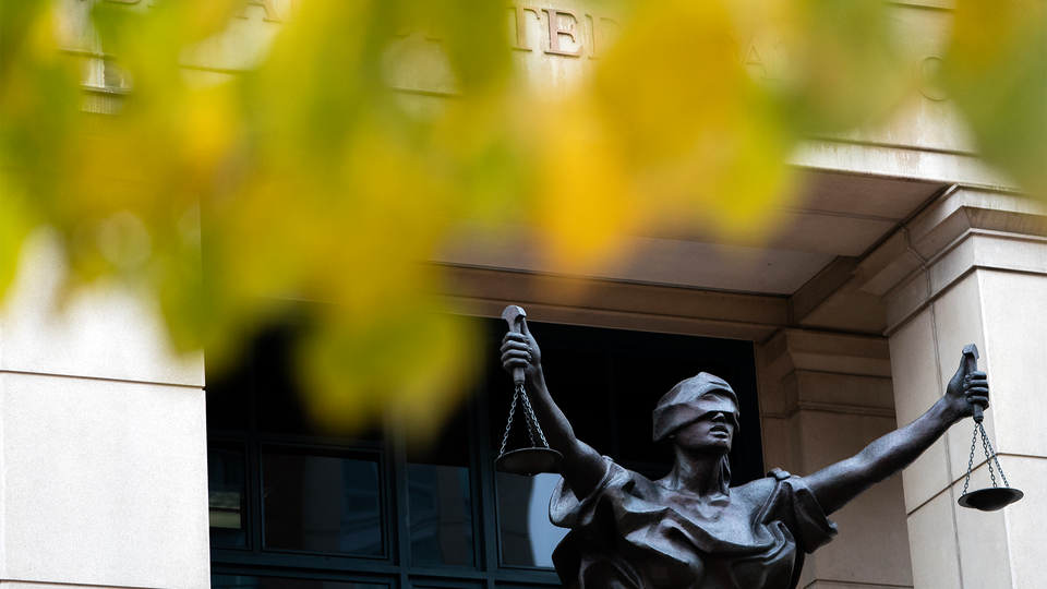 Photograph of a statue of Blind Justice outside the Albert V. Bryan United Sates Courthouse, autumn leaves in the foreground