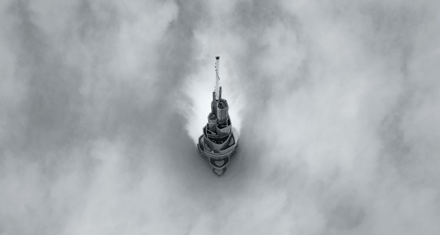 The top of a tall skyscraper is seen from above, piercing through low-lying clouds.