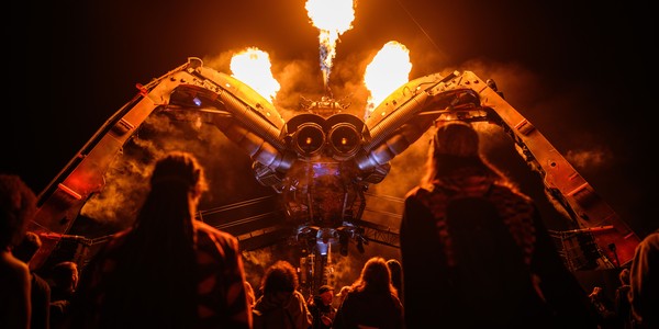 Fire erupts from the top of a large robotic spider sculpture as people look on below.
