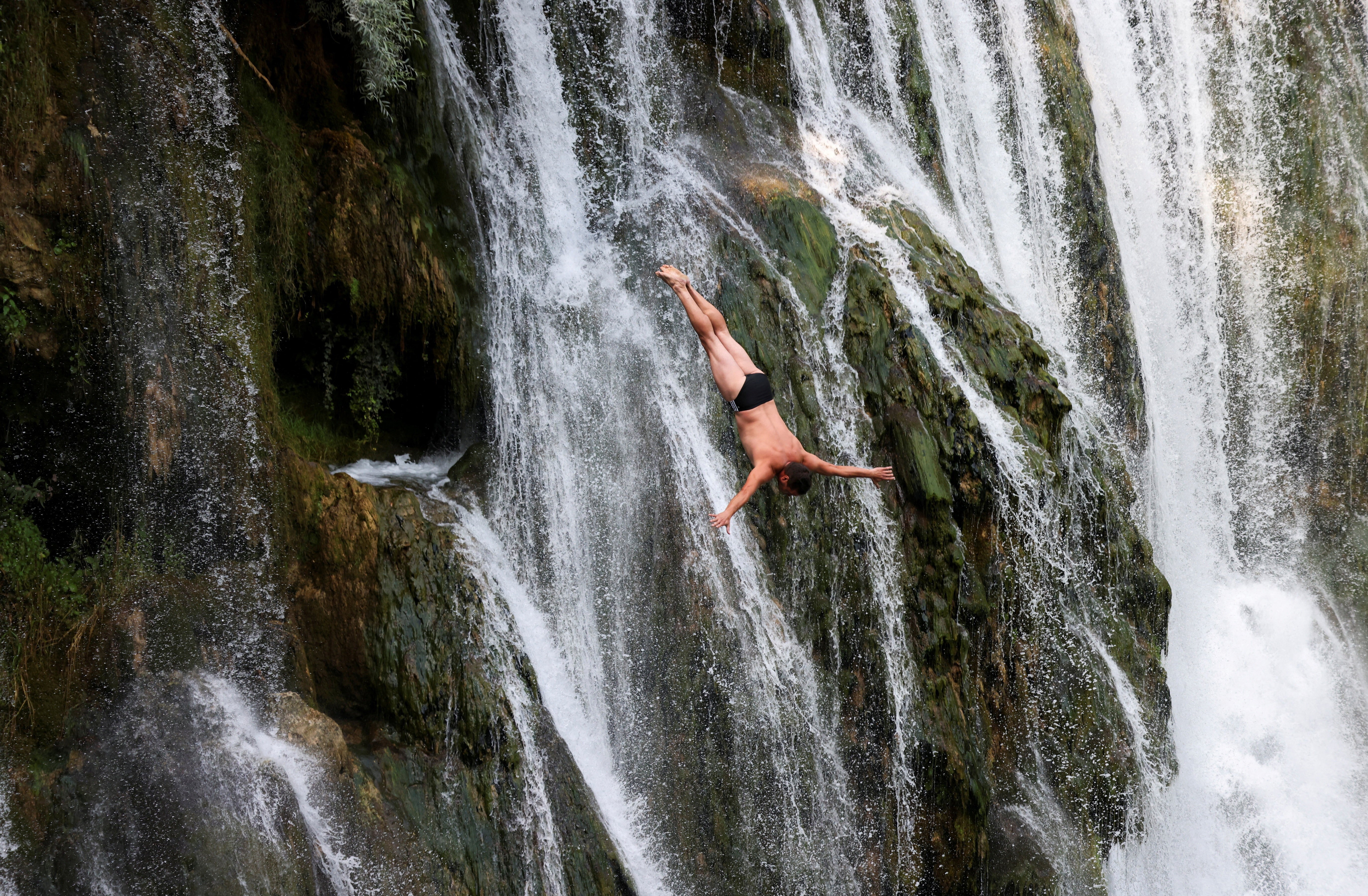 A driver plunges head-down toward an unseen pool, in front of a multi-channeled cliffside waterfall.