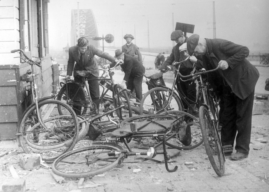 Several people examine a pile of abandoned bicycles near a bridge.