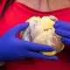 A woman wearing a red shirt and plastic gloves holds her own heart in front of her chest.