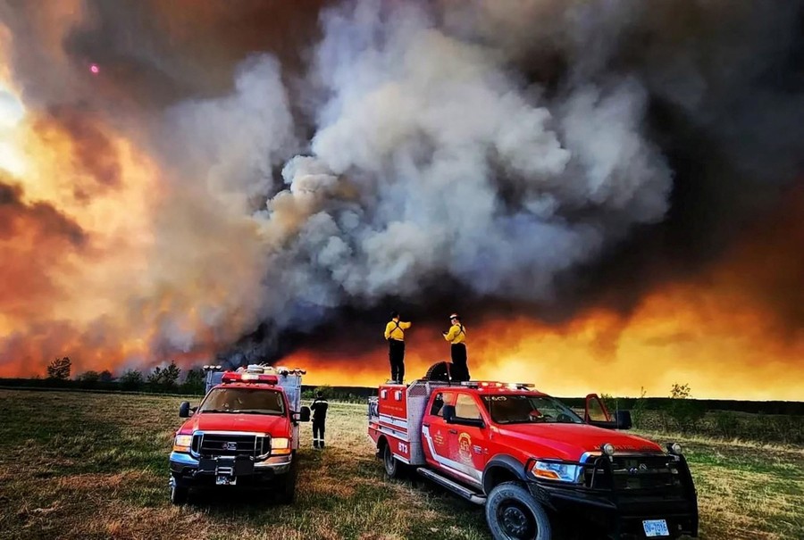 Two firefighters stand on top of a truck, looking toward a sky filled with smoke from a nearby forest fire.