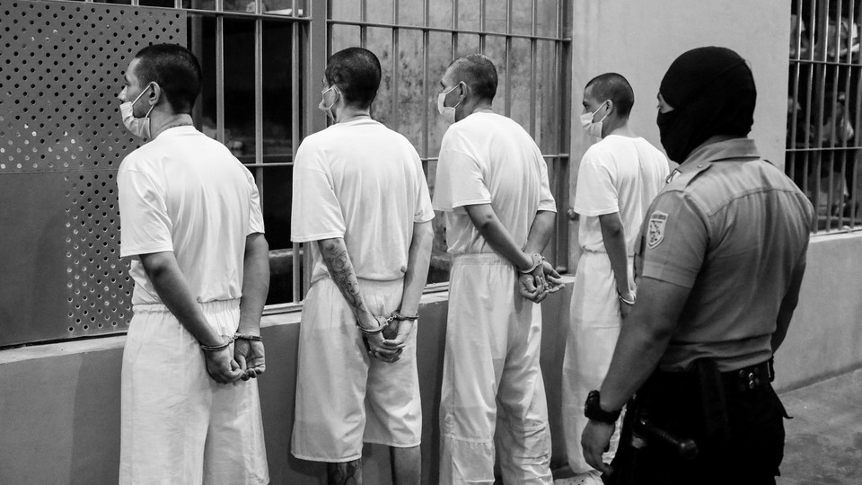Photograph of four handcuffed prisoners in El Salvador being watched from behind by a masked prison guard