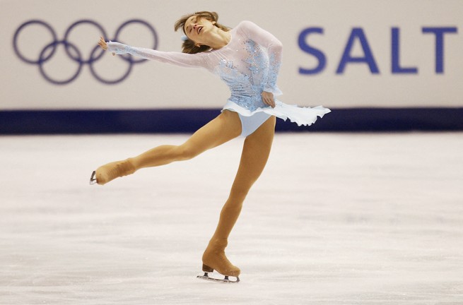 photo of woman in white short-skirted skating costume performing on ice rink with Olympic logo in background