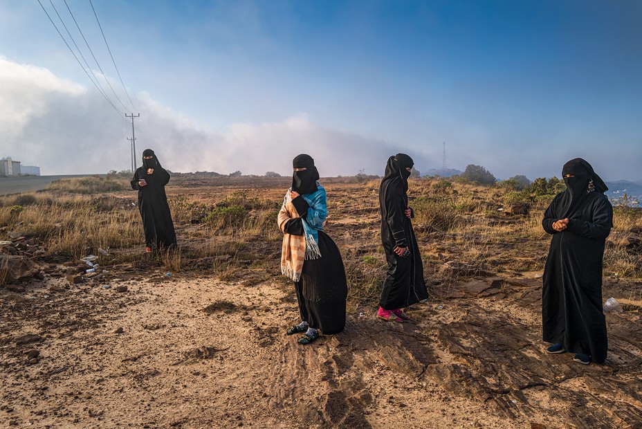 Four women in niqab and abayas stand separately in a rocky field 