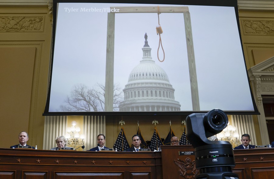 A photograph of a noose erected in front of the U.S. Capitol Building is projected on a screen above several people seated at the head of a meeting room.