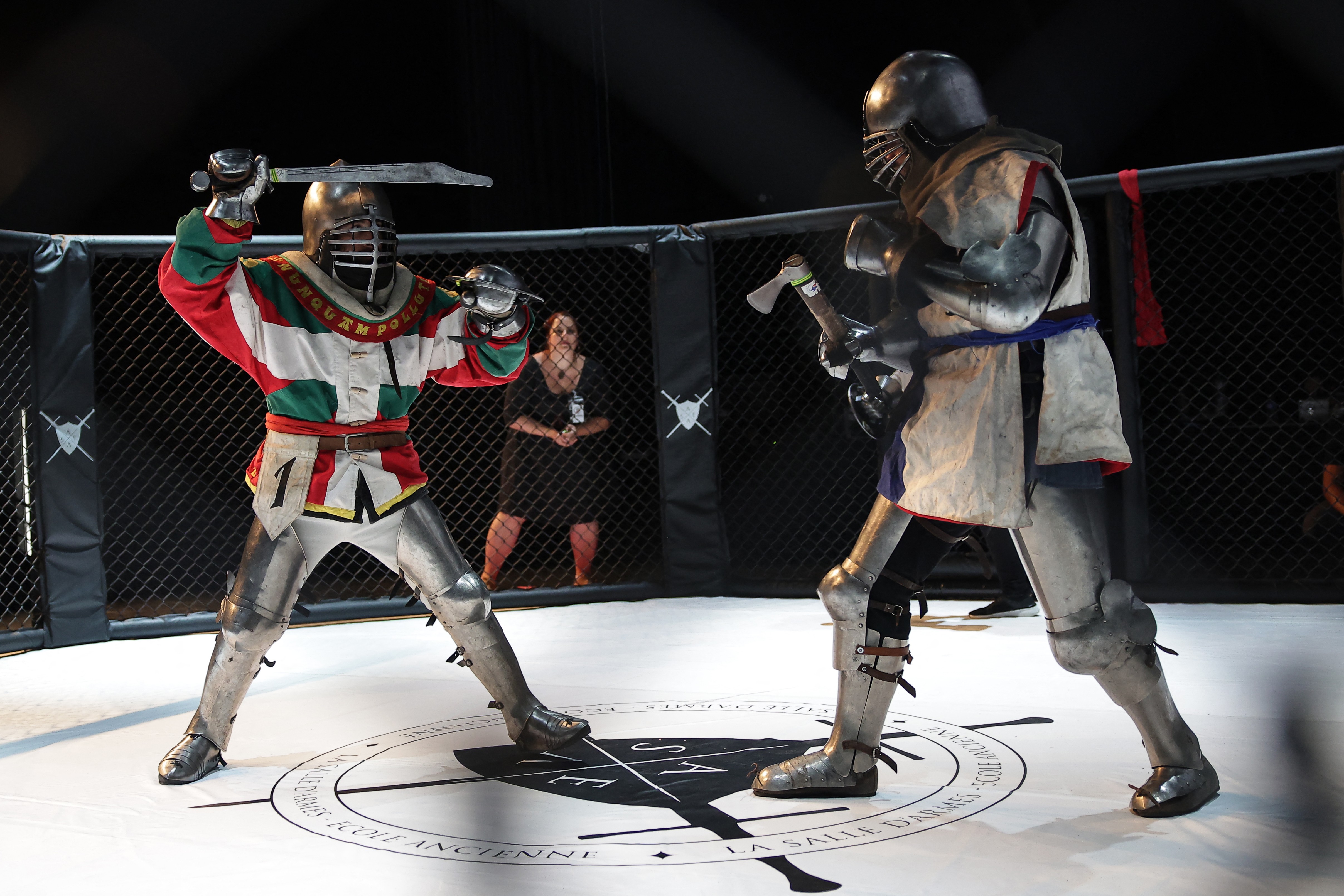 Two people, dressed in full medieval armor, face off inside a mixed martial arts fighting cage.